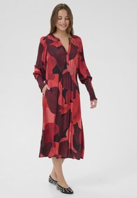 Rot und schwarz gemustertes Kleid mit V-Ausschnitt, langen Ärmeln und seitlichen Taschen; aus einem weichen, fließenden Stoff gefertigt. Schwarze Ballerinas mit Nieten.