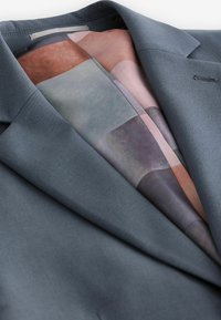 Grauer Blazer aus einem glatten Stoff, mit einem gemusterten Innenfutter in gedämpften Tönen von Rosa, Grau und Braun, das moderne Designelemente zeigt.