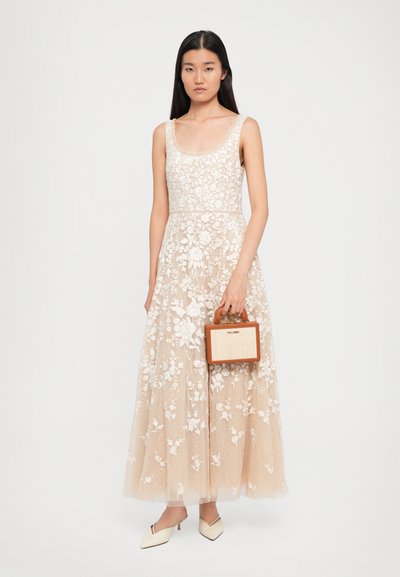 Femeie îmbrăcată într-o rochie maxi fără mâneci, cu dantelă florală bej, ținând o geantă de umăr rectangulară mică, în nuanțe bej și crem, asortată cu tocuri crem.
