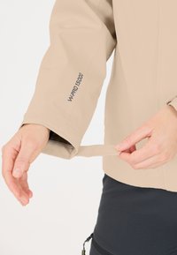 Beige vattentät jacka med justerbar manschett, tryckt "W-PRO 15000" på ärmen. Slät textur, figursydd design, visad i en handjusterande position.