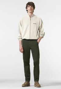 Helle beige Jacke mit Reißverschluss und schwarzem Text, kombiniert mit dunkelgrünen Hosen und beigen Stiefeln. Glatte Textur, sportliches Design. Schlichter Hintergrund.