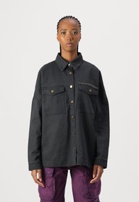 Giubbotto di jeans nero con colletto, chiusura con bottoni dorati, due tasche sul petto e un accento decorativo di borchie sulla tasca.