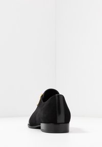 Chaussure en daim noir avec un bout arrondi, des coutures discrètes et un talon noir brillant. Elle possède des lacets dorés.