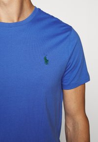 Modrá bavlněná tričko s krátkým rukávem a kulatým výstřihem; na levé straně hrudi má vyšitý zelený logo. Hladká textura.