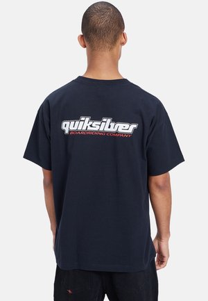 Personne portant un t-shirt noir Quiksilver avec le texte « BOARD RIDING COMPANY » sous le logo, debout de dos sur un fond clair uni.