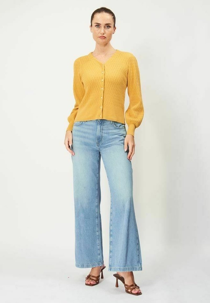 Senapsgul stickad kofta med V-ringning och knappstängning, i kombination med ljusblå vida jeans och bruna sandaletter med öppen tå.