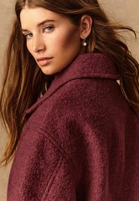 Manteau texturé bordeaux avec un grand col, en tissu doux et au coupe décontractée. Le mannequin porte une boucle d'oreille en forme de cœur.