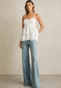 Kvinna i vit blommig spetslinne med tunna remmar och vida ljusblå jeans, stående mot en enkel beige vägg.