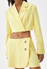 Cropped yellow blazer with notched lapels, paired with a matching mini skirt featuring a wrap design and two golden buttons. Smooth texture.