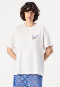 Balta medvilnė t-shirt su trumpomis rankovėmis, turinti spalvingą drugelių spaudą ant krūtinės. Drabužis yra laisvo, atsipalaidavusio kirpimo.