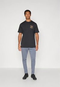 T-shirt preta de mangas curtas com logótipo dourado no peito, combinada com calças de fato de treino cinza justas e ténis pretos. Fundo simples.