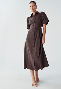 Hnědé šaty na knoflíky s balónovými rukávy, opasek na zavázání v pase a splývavý střih. Měkká látka s hladkým povrchem, délka po kotníky.