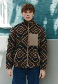 Brown fleece jacket with geometric patterns in dark brown, cream, blue, and green. Features a beige chest pocket and zip closure.