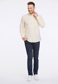 Ljusgul, långärmad knäppskjorta med bröstficka, matchad med mörkblå jeans och vita sneakers, på en stående modell.