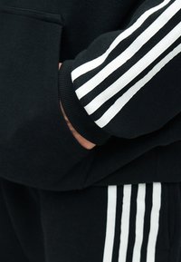 Persona con abbigliamento sportivo nero con dettagli bianchi a tre strisce su manica e pantaloni, mano infilata nella tasca anteriore.