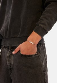 Stříbrný náramek na zápěstí, se žlutým obdélníkovým přívěskem, nošený s černou mikinou a roztrhanými černými džínami.