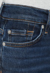 Calças de ganga azul escuro com costuras laranja, um pequeno bolso frontal e detalhes em botões metálicos na cintura. Textura suave.