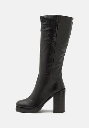 Botte en cuir noire montant à mi-mollet, avec bout arrondi, talon épais et carré, et détail de couture subtile sur un fond clair uni.