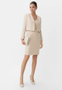 Beiges, enganliegendes Kleid mit einer kurzen Jacke. Das Kleid hat einen V-Ausschnitt und eine glatte Textur. Die Accessoires umfassen eine leichte Clutch und spitze Absätze.