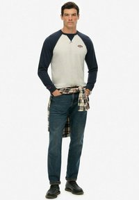 Camisa de béisbol de manga larga en crema y azul marino, combinada con una camisa de franela a cuadros atada a la cintura, jeans azules y zapatos negros.
