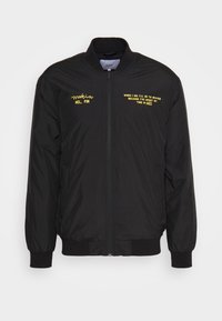 Giacca bomber nera realizzata in tessuto liscio. Presenta testo e logo gialli sul petto, chiusura zip frontale, polsini e orlo a costine.