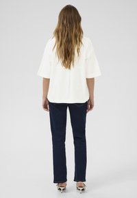 White textured blouse with loose sleeves, paired with dark blue straight-leg jeans and strappy heels. Hair falls in loose waves.