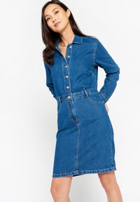 Modra jeans obleka z dolgimi rokavi, gumbom na sprednji strani, pasom in žepi. Ima ravno krojenje in subtilne detajle pri šivanju.