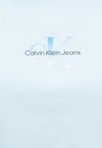 Calvin Klein Jeans-logo med store "ck"-initialer i gradient blått og hvitt på en ensfarget lys blå stoffbakgrunn.
