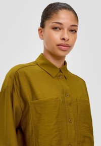 Olijfgroene shirt met knoopsluiting aan de voorkant, kraag en twee borstzakken. Textuurstof met een losse pasvorm en zichtbare stikseldetails.