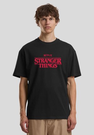 Tricou negru din bumbac, cu un logo îndrăzneț „STRANGER THINGS” roșu, cu „NETFLIX” deasupra, cu mâneci scurte, croială lejeră și decolteu rotund.