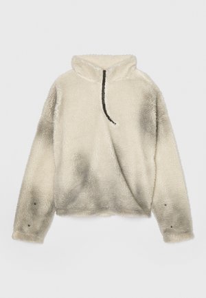Udune, kreemjas värvi poolkõrge kaelusega pullover, millel on pool-zip sulgemine, tekstuuriga pind ja mustad aktsendid kaeluse ja varrukate ääres.