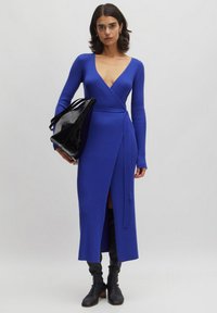 Langes, tiefblaues, geripptes Strickkleid mit V-Ausschnitt und Wickeldesign. Mit langen Ärmeln und einem seitlichen Schlitz. Begleitet von einer schwarzen, glänzenden Tasche.