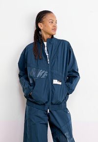 Tumši zils Nike vēja jaku ar logotipa detaļām, priekšējām kabatām un augstu apkakli. Iespējām brīvu piegriezumu un vieglu audumu.