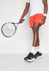 Κόκκινες σορτς, λευκή μπλούζα, μαύρα παπούτσια Nike και μια ρακέτα τένις με μπλε λεπτομέρειες. Οι σορτς έχουν λεπτομέρεια με λογότυπο και είναι φτιαγμένες από ελαφρύ υλικό.