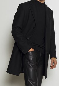 Homme portant un manteau noir à double boutonnage, un pull à col roulé noir et un pantalon en cuir noir, la main dans la poche.