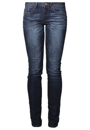 Frau trägt einen grauen Cardigan, ein weißes Top, blaue Jeans, Sneakers und trägt einen schwarzen Rucksack über einer Schulter, steht vor einem schlichten Hintergrund.