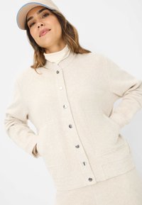 Beige Wollmischung Jacke mit Knopfverschluss, zwei vorderen Taschen und gerippten Bündchen. Der Stoff hat eine weiche Textur und ein dezentes Muster.