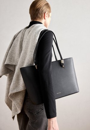 Personne aux cheveux courts portant un haut noir et un pantalon gris, porte un grand sac fourre-tout noir sur l’épaule avec un pull gris drapé sur le dos.