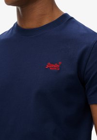 Primer plano de una persona que lleva una camiseta de cuello redondo azul marino con un pequeño logo bordado en rojo de "SuperDry" en el pecho izquierdo.