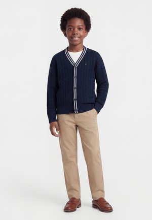 Junge lächelt, trägt einen navyblauen, geknöpften Cardigan, weißes Unterhemd, beige Hose und braune Loafers, steht mit einer Hand in der Tasche.