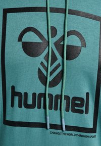Türkiser Hoodie mit schwarzem Logo, das abstrakte Formen zeigt, verstellbarem Kordelzug mit blauen Enden und dem Text "Verändere die Welt durch Sport."