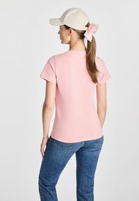 Różowa koszulka z krótkim rękawem o dopasowanym fasonie, w połączeniu z niebieskimi dżinsami. Na głowie jest jasnobeżowa czapka, a włosy związane są różową kokardą.