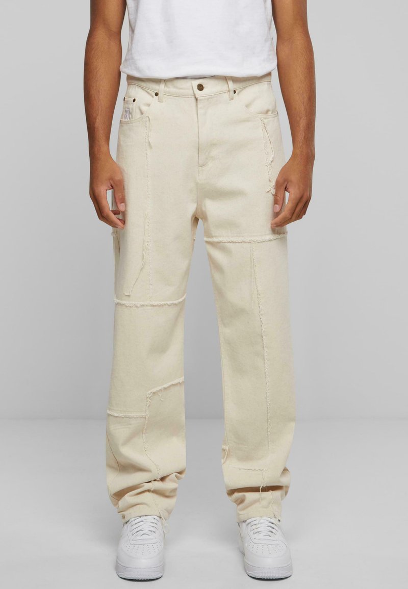Beige, brede bukser laget av denim, med synlige sømmer, revne detaljer og to sidelommer. Brukt med hvite joggesko.