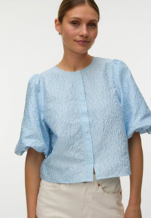 Femme portant un chemisier bleu clair texturé à manches courtes bouffantes et un pantalon crème sur un fond uni.