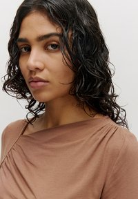 Braun drapierter Oberteil mit breitem Ausschnitt und weichem Ripptextur. Das Model hat lockiges, dunkles Haar und minimales Makeup, wobei der Fokus auf dem Kleidungsstück liegt.