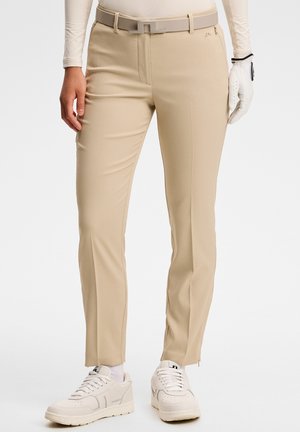 Person iført beige tætsiddende bukser med matchende bælte, hvid golfhandske på højre hånd, hvide sneakers og hvide sokker på en ensfarvet baggrund.