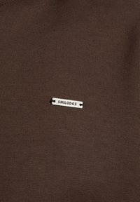 Hnědá látka s malou obdélníkovou stříbrnou kovovou nálepkou přišitou, vygravírovanou se slovem "SMILODOX".