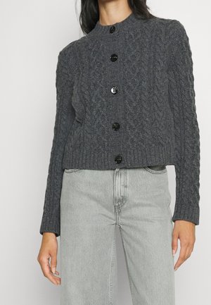 Kvinde iført en mørkegrå kabelstrikket cardigan med knapper og lysegrå højttaljede jeans, vist fra nakke til hofter.