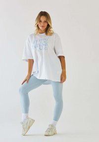 Biała, oversize'owa koszulka z niebieskim napisem, zestawiona z jasnoniebieskimi legginsami. Modelka w białych skarpetkach i beżowych sneakersach, stojąca w pozie.