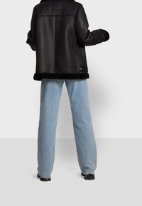 Persona con chaqueta negra de borrego y pantalones vaqueros anchos azul claro, de pie de espaldas contra un fondo liso.